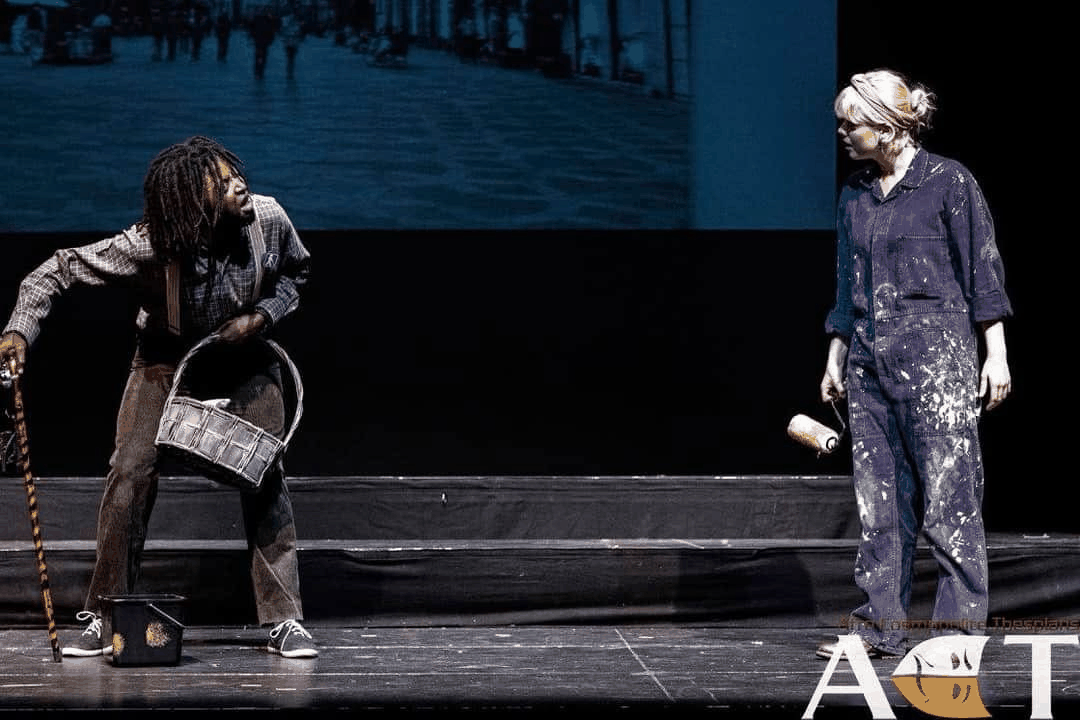 Launcelot in a comic stage still from The Merchant of Venice holding a bucket and stick while arguing with another character.